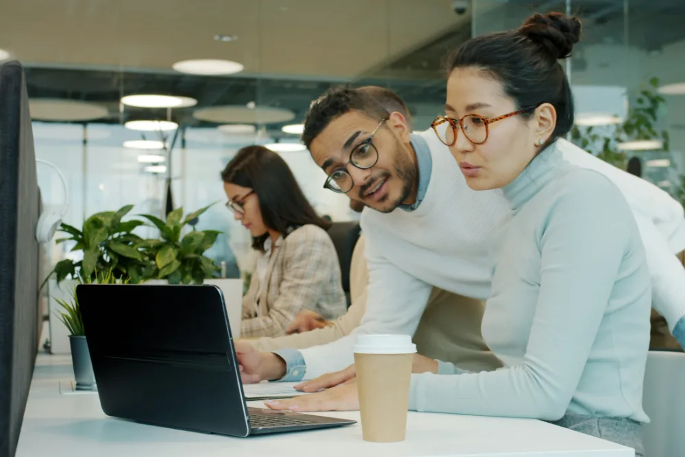Two colleagues reviewing something together on a laptop in an open-plan office.
