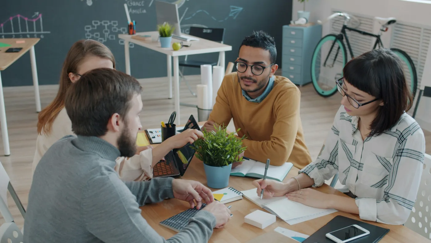 A team of four working together at a shared desk with laptops and notebooks.